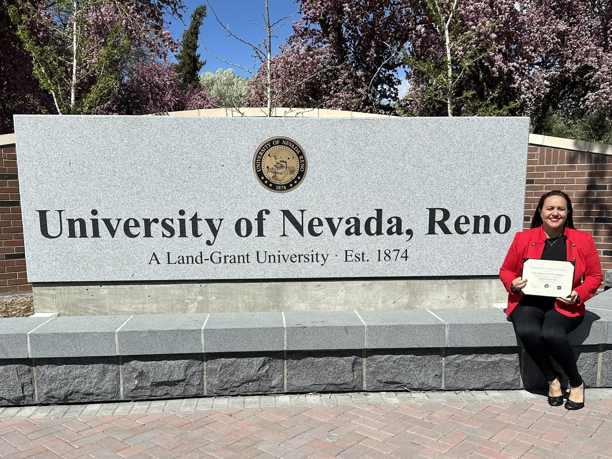 Woman holds diploma in front of university sign.